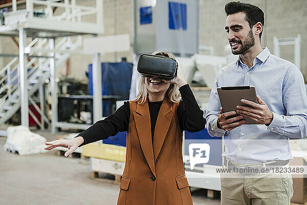 Happy businessman holding tablet PC and standing by colleague wearing virtual reality simulator