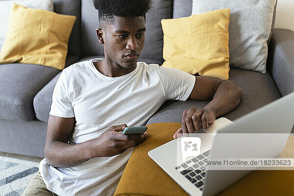 Mann mit Smartphone und Laptop sitzt zu Hause neben dem Sofa