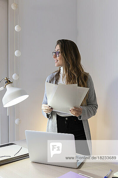 Smiling young woman standing with documents at desk