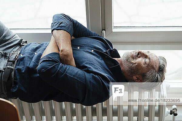 Mature man lying on radiator by window at home