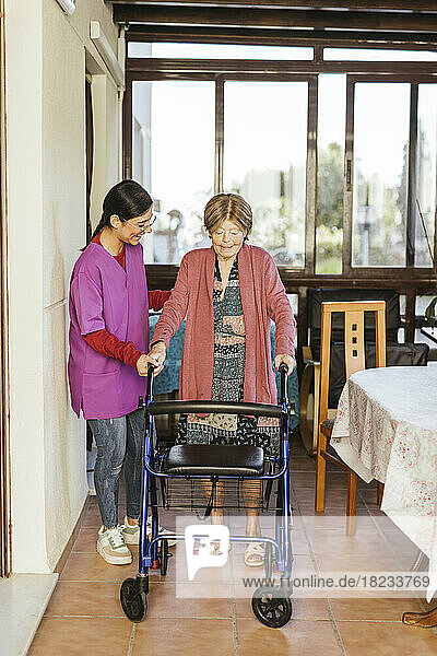 Lächelnde junge Pflegekraft hilft einer älteren Frau  zu Hause mit einem Gehhilfe zu gehen