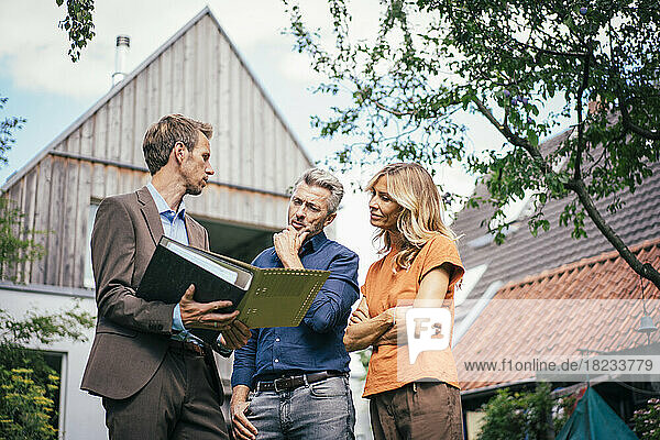 Couple discussing over documents with real estate agent in front of house