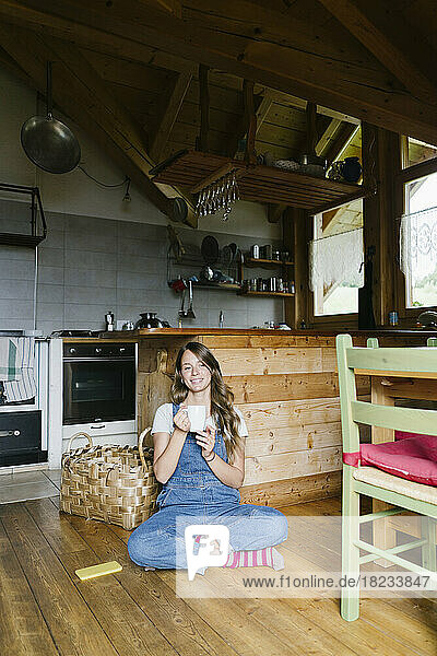 Glückliche Frau mit Kaffeetasse sitzt auf dem Boden in der Küche