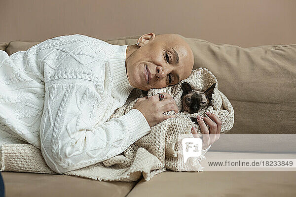 Bald woman embracing Sphynx cat wrapped in blanket on sofa