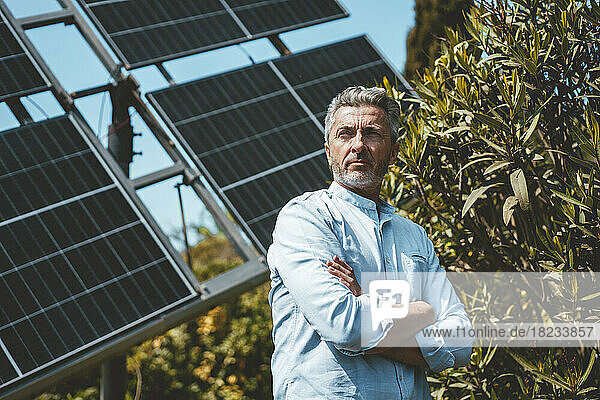 Thoughtful man with arms crossed in front of solar panels on sunny day
