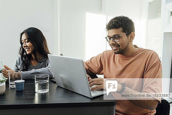 Smiling man studying through laptop sitting by woman at table in living room