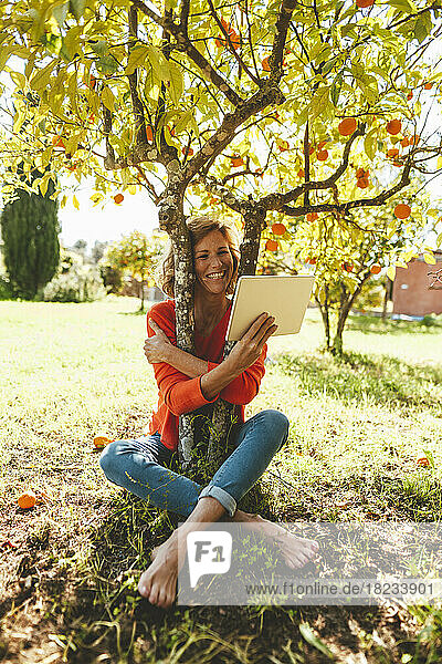 Happy mature woman hugging orange fruit tree and using tablet PC