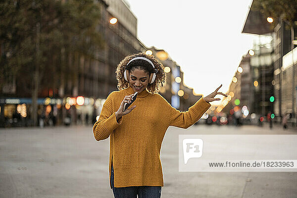 Lächelnde junge Frau hält ihr Mobiltelefon in der Hand  genießt Musik über kabellose Kopfhörer und tanzt auf dem Fußweg