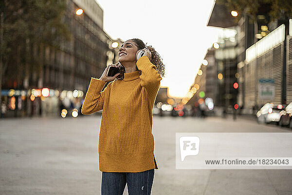 Glückliche Frau hält ihr Mobiltelefon in der Hand und genießt Musik über kabellose Kopfhörer auf dem Fußweg