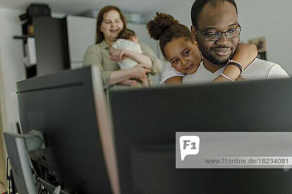 Smiling man working through computer with daughter embracing from behind