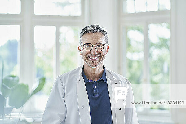 Happy mature doctor wearing eyeglasses standing in medical practice