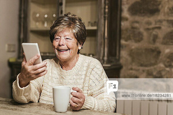 Glückliche ältere Frau  die ihr Smartphone benutzt und zu Hause mit einer Kaffeetasse sitzt