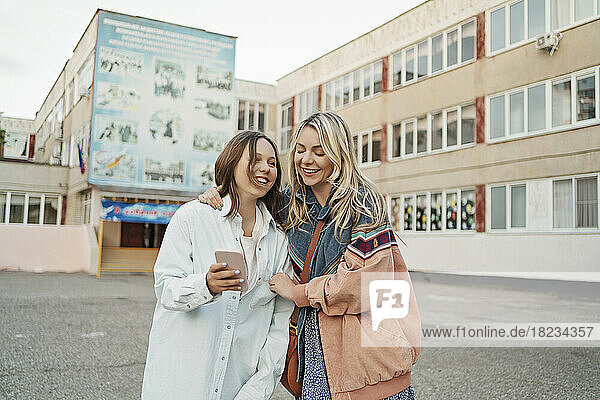 Happy mother and daughter sharing smart phone standing on schoolyard