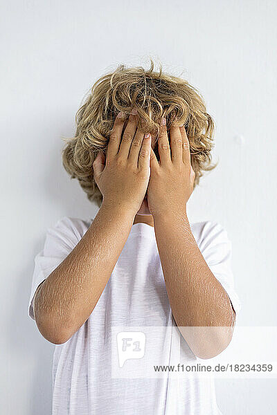 Shy boy covering face with hands against white background