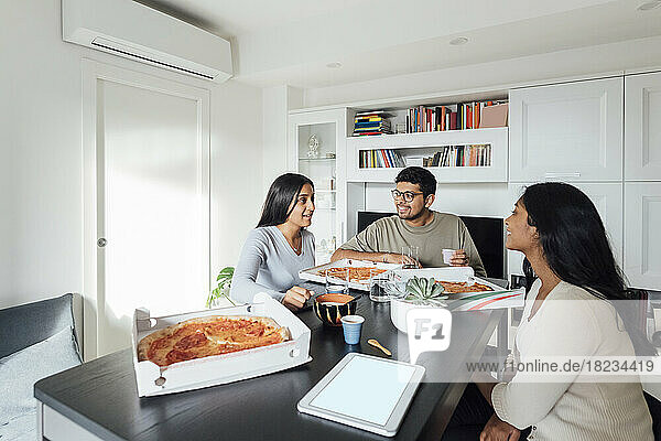 Glückliche Frauen und Männer diskutieren und essen Pizza am Esstisch im Wohnzimmer