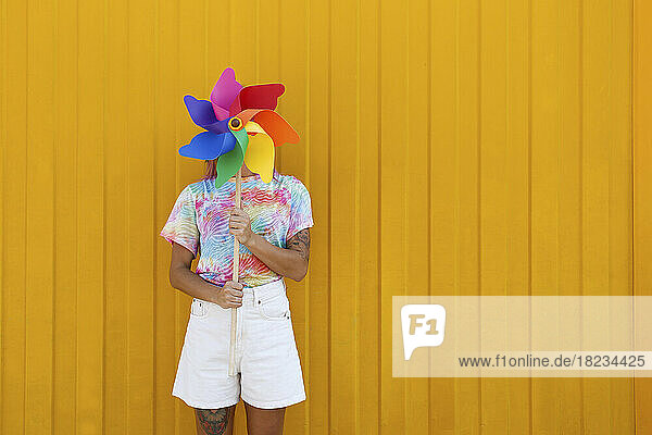 Young woman covering face with rainbow colored pinwheel toy in front of yellow wall