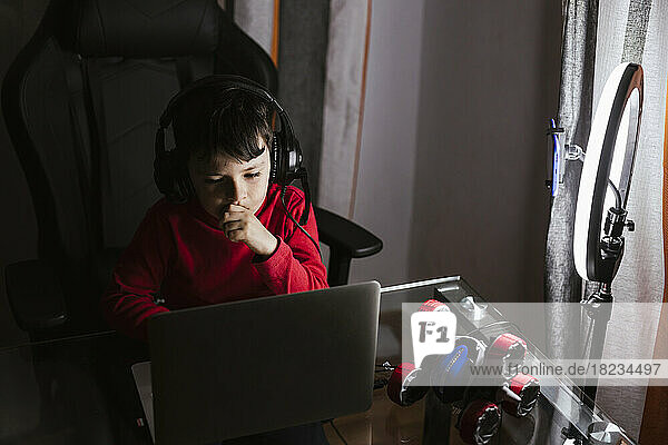 Boy wearing headphones using laptop sitting by mobile phone in illuminated ring light