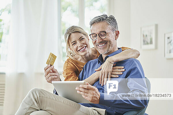 Happy woman embracing man with credit card doing online shopping through tablet PC at home