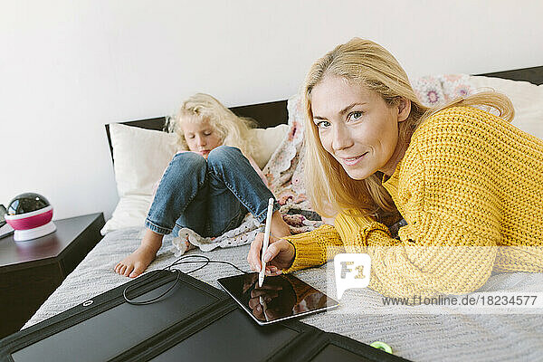 Mother holding digitized pen with tablet PC by daughter on bed