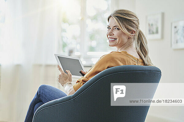 Happy woman with blond hair holding tablet PC in armchair