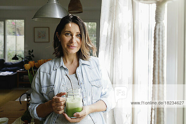 Smiling woman with glass of green smoothie standing at home