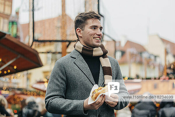 Happy young man with hot dog standing at Christmas market