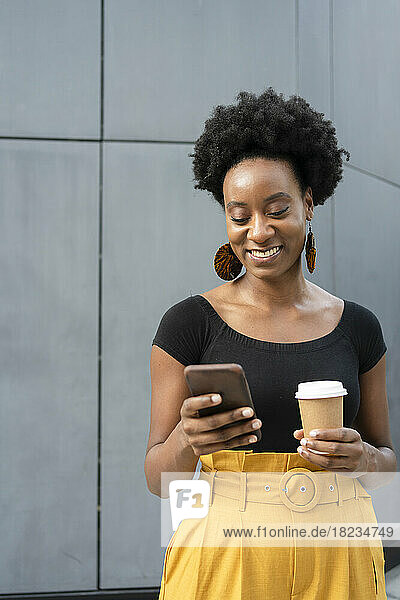 Smiling woman using mobile phone and holding disposable cup in front of wall
