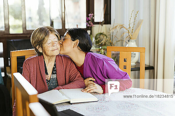 Caregiver kissing senior woman sitting at table in home