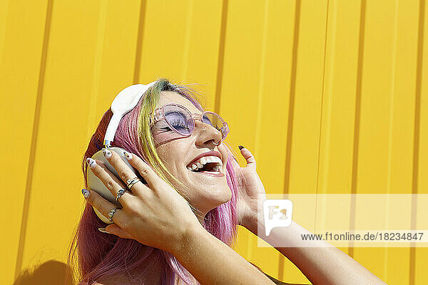 Happy woman wearing sunglasses enjoying music listening through wireless headphones in front of yellow wall