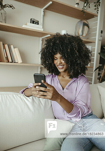 Smiling Afro woman using smart phone sitting on sofa at home