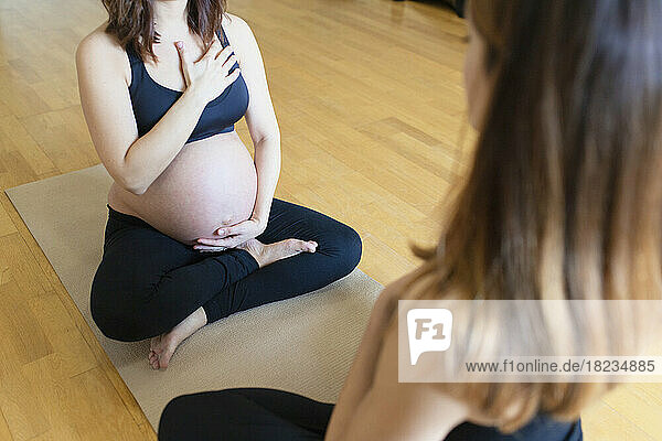 Pregnant woman keeping hand on stomach and chest practicing yoga with instructor in studio