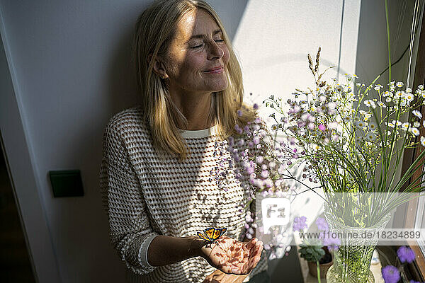 Mature woman standing with butterfly on hand and sunlight over face at home