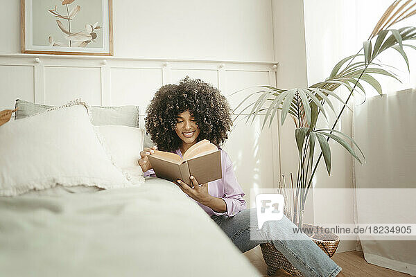 Smiling woman reading book sitting by bed in bedroom