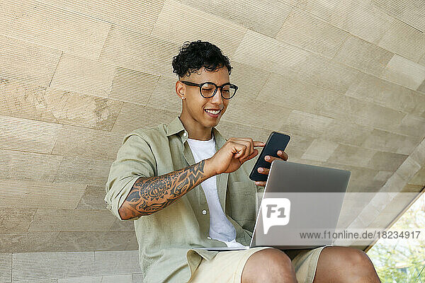 Smiling freelancer using smart phone sitting with laptop
