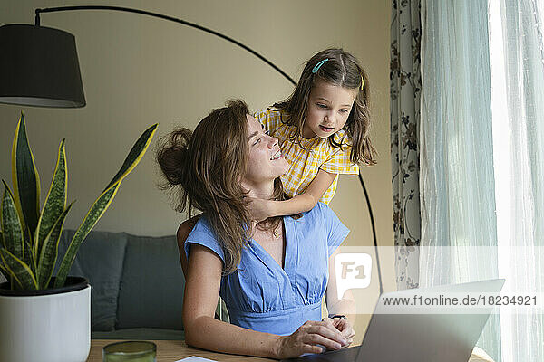 Süße Tochter umarmt Mutter  die mit Laptop am Schreibtisch im Heimbüro sitzt