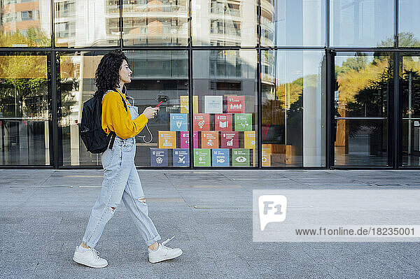 Young woman with backpack holding mobile phone and walking on footpath