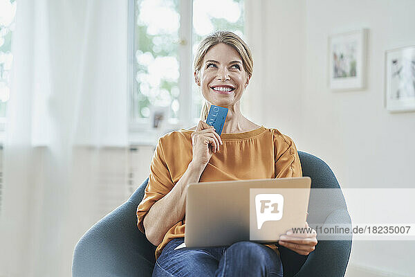 Thoughtful smiling woman holding credit card and laptop on armchair at home