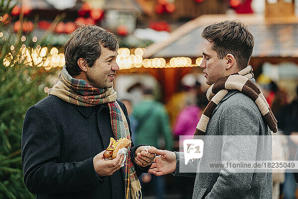 Smiling father holding hot dog and talking with son standing at Christmas market