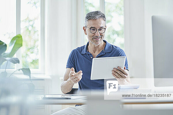 Smiling mature doctor gesturing and talking on video call through tablet PC