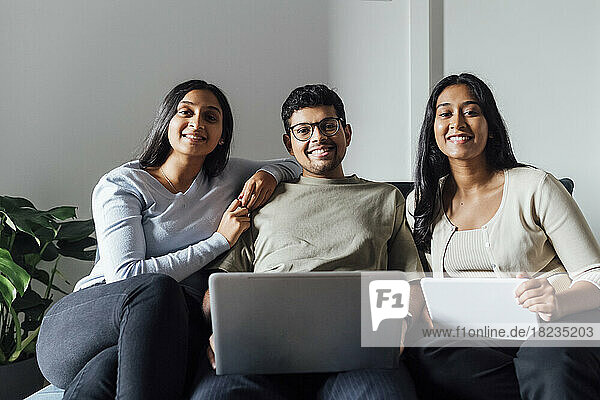 Smiling women and man with laptop and digital tablet sitting on sofa