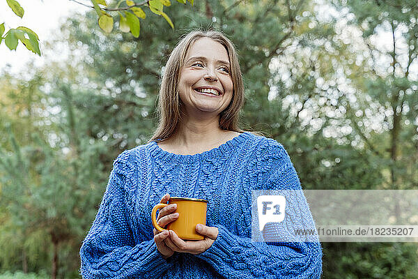 Lächelnde Frau im blauen Pullover mit Kaffeetasse und Tagträumen im Freien