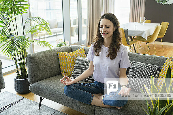 Lächelnde Frau meditiert zu Hause auf dem Sofa im Wohnzimmer