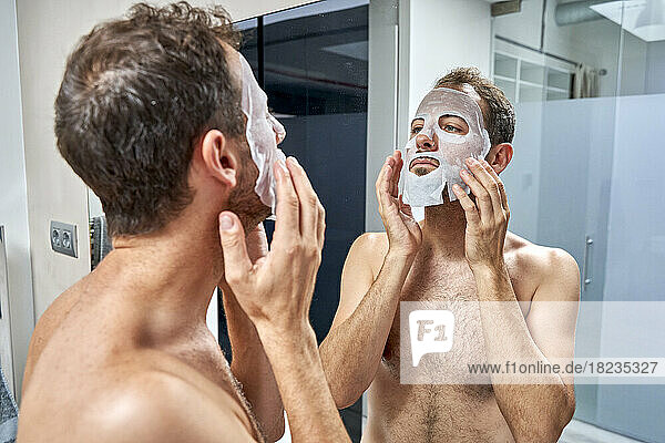 Man applying sheet mask on face looking in mirror at home