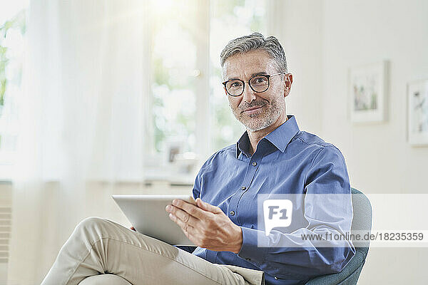 Lächelnder Geschäftsmann hält Tablet-PC im Sessel zu Hause