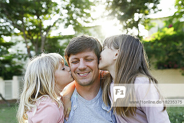 Lächelnder Vater mit Töchtern  die ihn im Park auf die Wange küssen