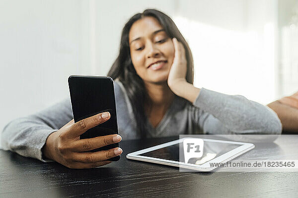 Smiling woman using smart phone sitting with tablet PC on table at home