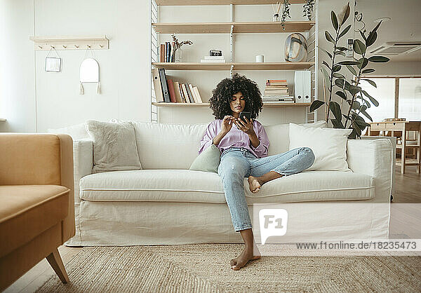 Relaxed Afro woman using smart phone sitting on sofa at home
