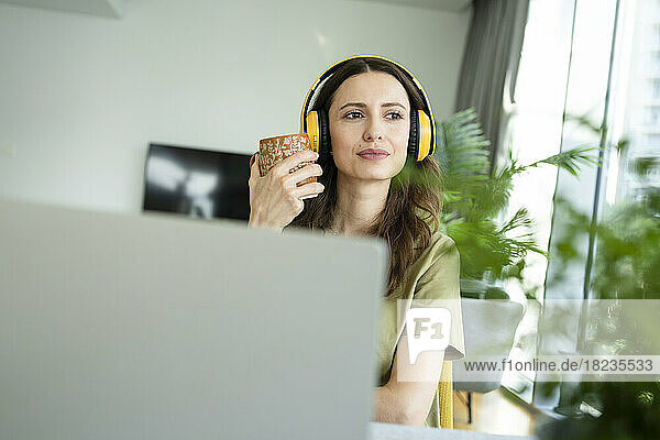 Geschäftsfrau trägt kabellose Kopfhörer und hält eine Kaffeetasse in der Hand und blickt zu Hause durch das Fenster