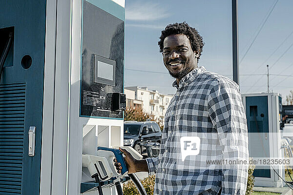 Smiling man standing at car charging station on sunny day
