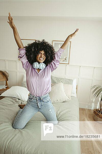 Afro woman with arms raised dancing on bed in bedroom